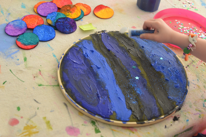 Réalisez un système solaire avec des rondelles de coton, de l'aquarelle, de l'acrylique, du tissu et un cerceau à broder.  Une merveilleuse activité artistique ouverte pour célébrer le jour de la Terre!