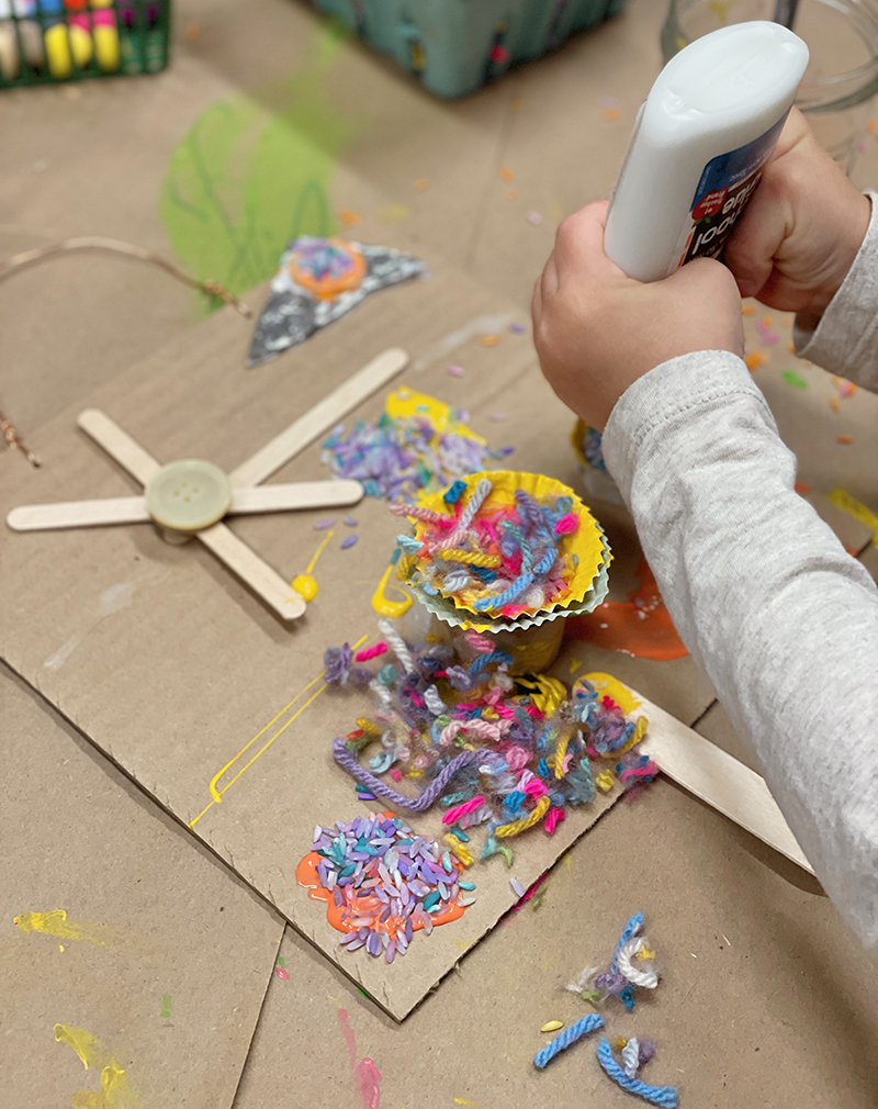 Enfant utilisant de la colle pour embellir son collage recyclé avec des morceaux de fil, du riz teint et des doublures de cupcake.